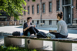 Two women are sitting on a bench outside the campus talking, one has a document in her hand.